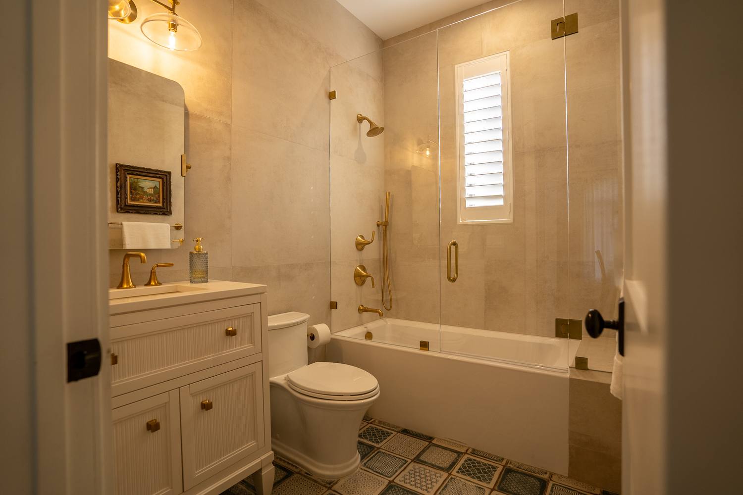 A bath with a patterned tile floor, a white vanity and brass fittings, a tub behind glass