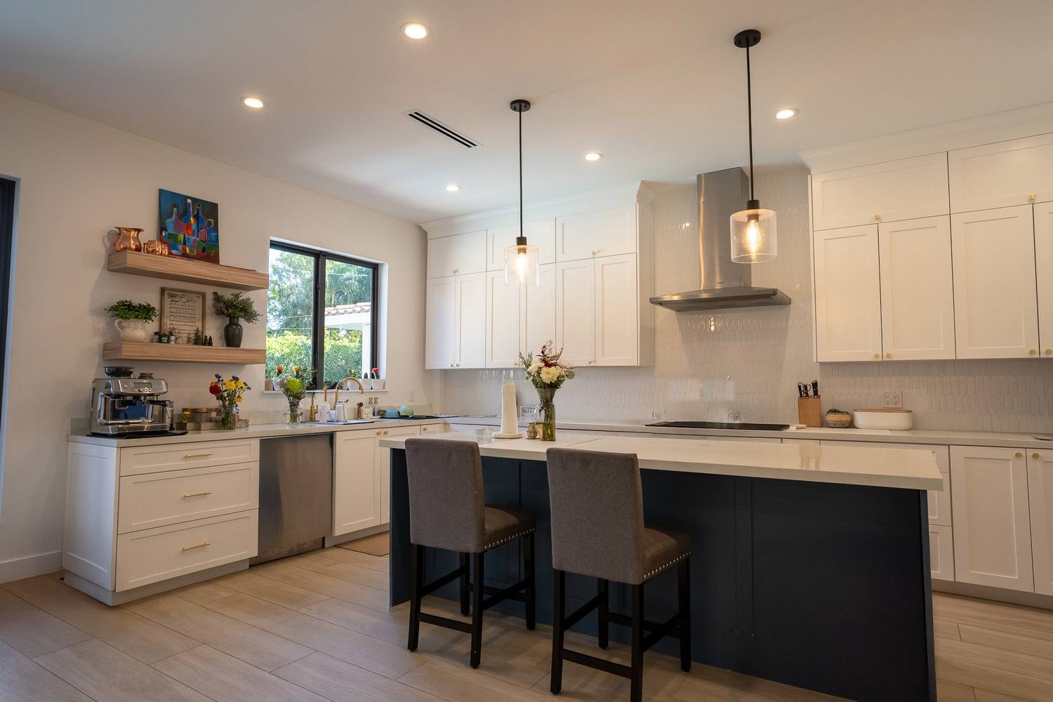 A kitchen with white shaker cabinets, a stainless hood and a navy island with two stools