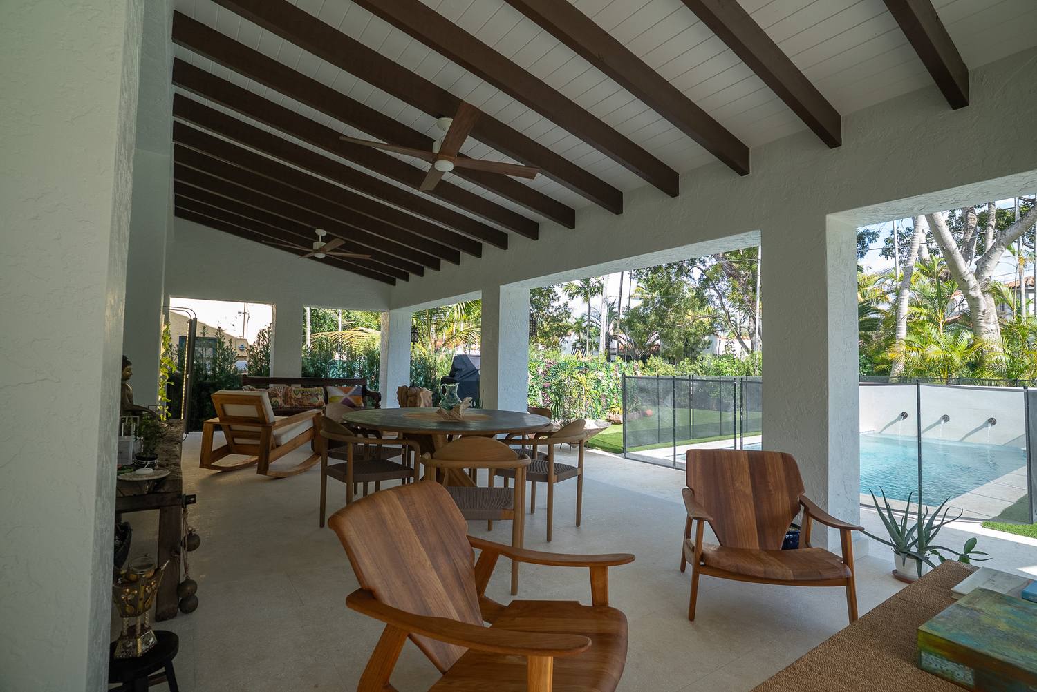 A covered terrace with wood beams and ceiling fans, wooden chairs, the pool and garden beyond