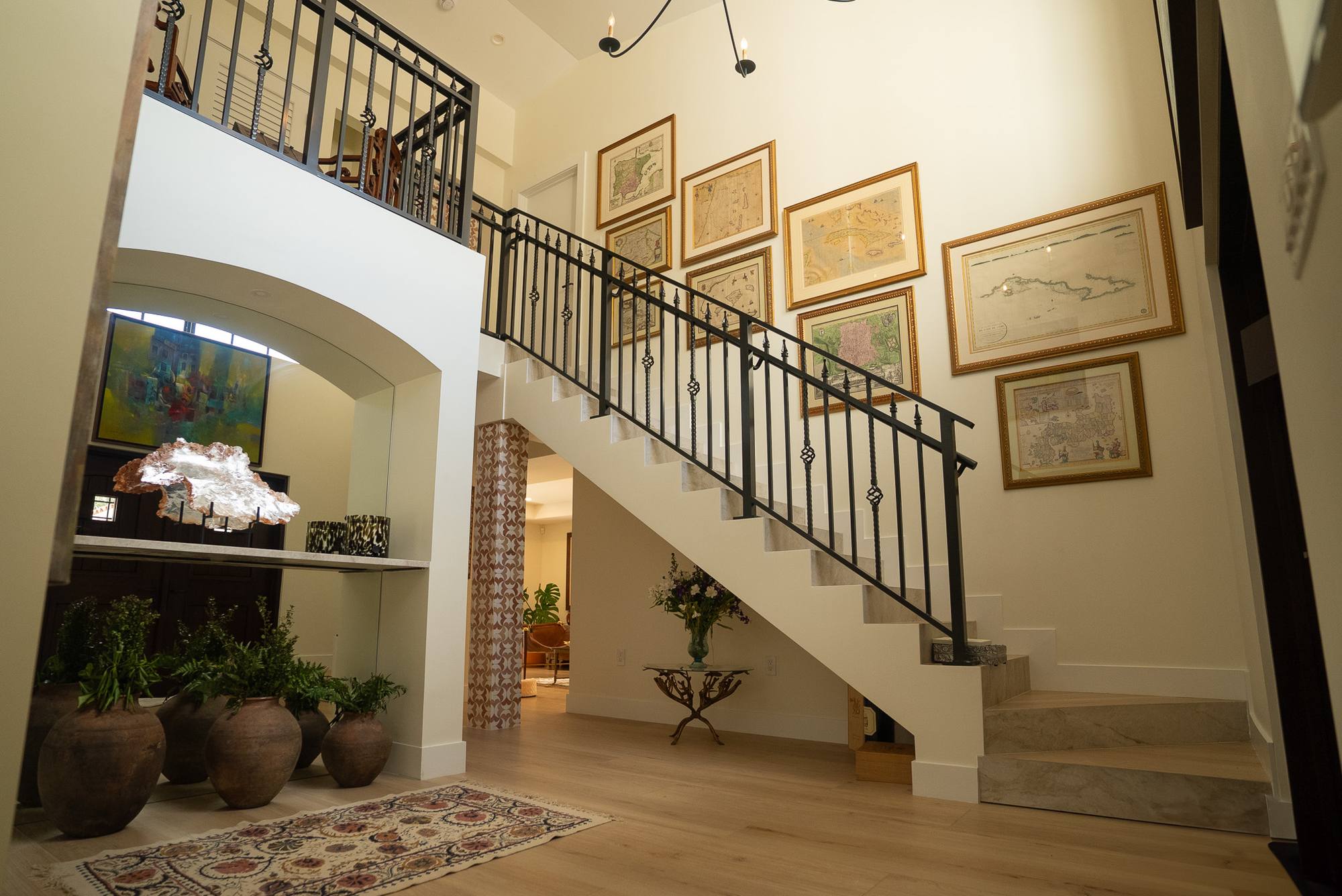 A double-height stair hall with an iron rail, framed maps on the wall and potted plants in an arched niche