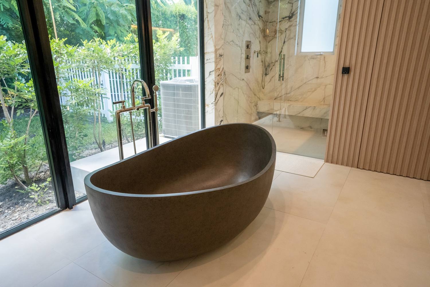 Freestanding dark stone tub by floor-to-ceiling glass looking onto the garden