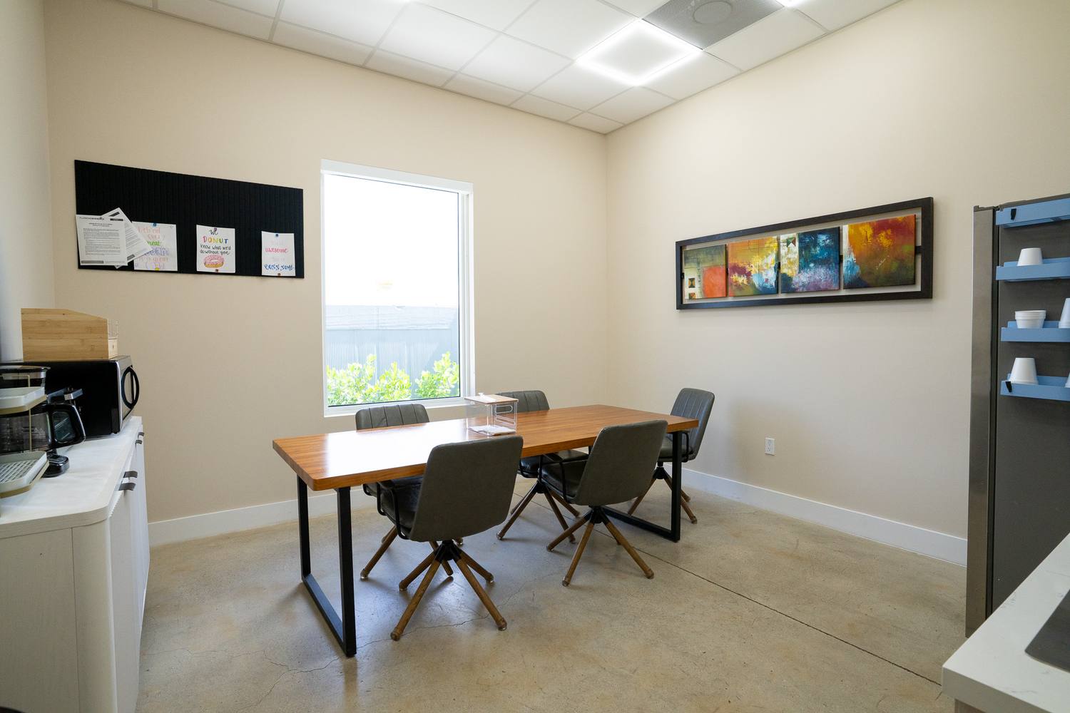 A break room with a wood table, grey chairs and a long painting on the wall