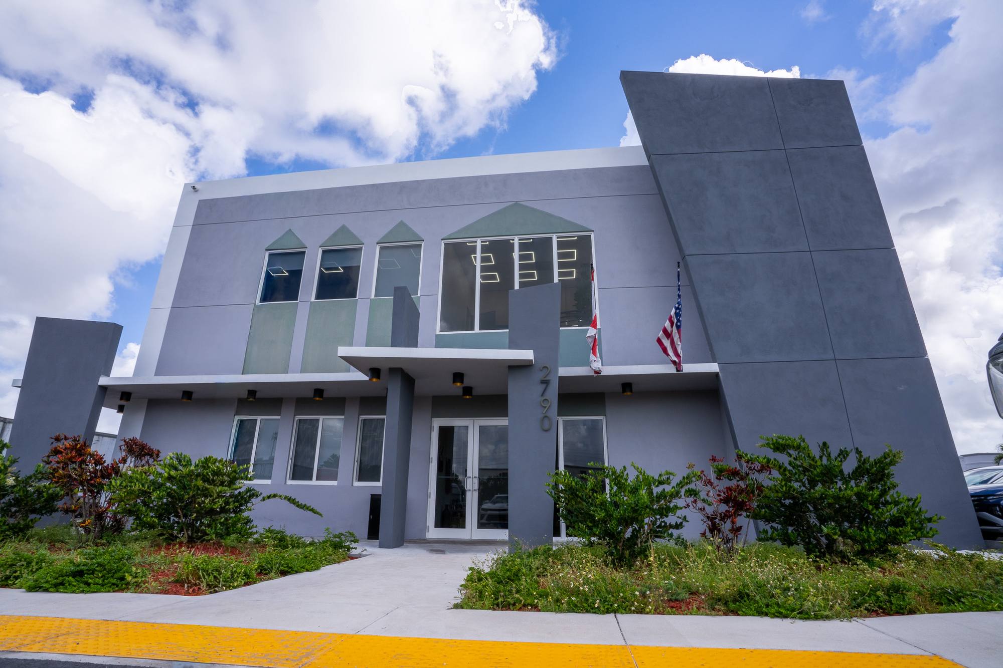 The office building's front entry under a flat canopy, grey stucco with angled wing walls and planted beds, Opa-locka